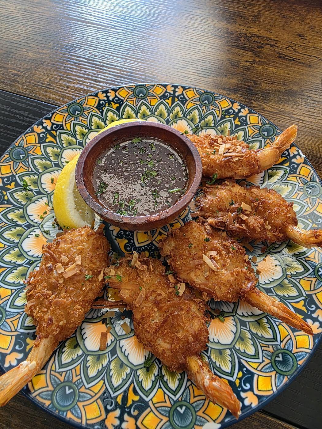 Coconut shrimp arranged on a patterned plate with a pot of dipping sauce and a wedge of lemon