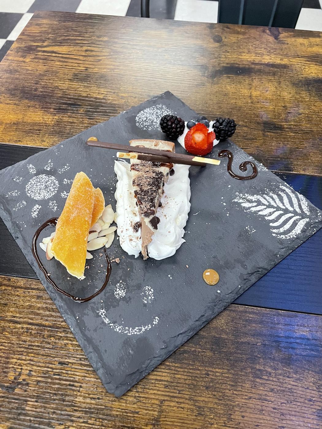 A dessert plated on a dark serving board with cream, berries and chocolate work